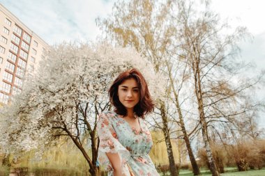 Portrait of charming pretty woman dressed flowery dress posing near apple cherry tree blossoms blooming flowers in the garden park in early spring nature. Fashion, girl model with black hair
