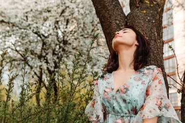 Portrait of charming pretty woman dressed flowery dress posing near apple cherry tree blossoms blooming flowers in the garden park in early spring nature. Fashion, girl model with black hair