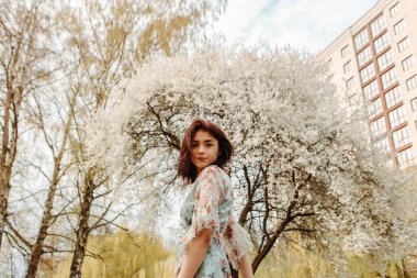 Portrait of charming pretty woman dressed flowery dress posing near apple cherry tree blossoms blooming flowers in the garden park in early spring nature. Fashion, girl model with black hair