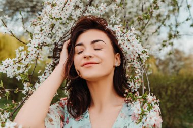 Portrait of charming pretty woman dressed flowery dress posing near apple cherry tree blossoms blooming flowers in the garden park in early spring nature. Fashion, girl model with black hair