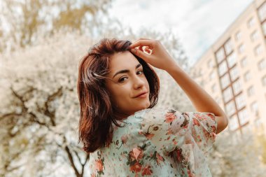 Portrait of charming pretty woman dressed flowery dress posing near apple cherry tree blossoms blooming flowers in the garden park in early spring nature. Fashion, girl model with black hair