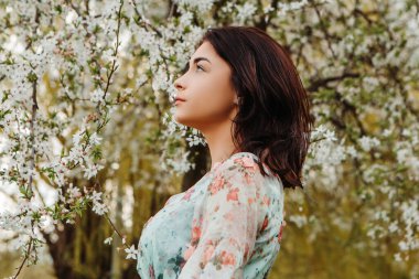 Profile pose looking to side woman dressed flowery dress posing near apple cherry tree blossoms blooming flowers in the garden park in early spring nature. Fashion, girl model with black hair