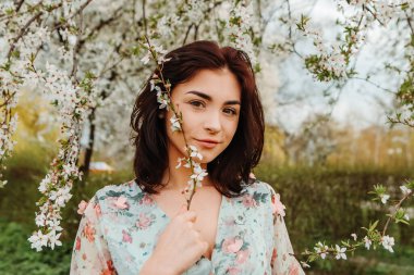 Portrait of charming pretty woman dressed flowery dress posing near apple cherry tree blossoms blooming flowers in the garden park in early spring nature. Fashion, girl model with black hair