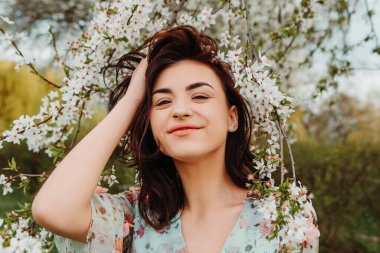 Portrait of charming pretty woman dressed flowery dress posing near apple cherry tree blossoms blooming flowers in the garden park in early spring nature. Fashion, girl model with black hair