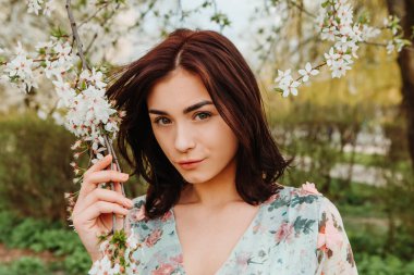 Portrait of charming pretty woman dressed flowery dress posing near apple cherry tree blossoms blooming flowers in the garden park in early spring nature. Fashion, girl model with black hair