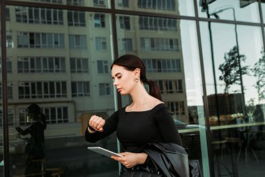 Pretty brunette caucasian woman dressed leather suit with shoulder bag stand outside hold tablet pc, looking at swart wrist wireless watch fitness bracelet, checking time, waiting in a hurry, punctual