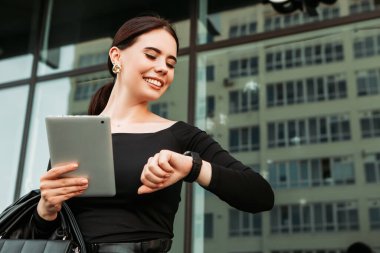 Pretty brunette caucasian woman dressed leather suit with shoulder bag stand outside hold tablet pc, looking at swart wrist wireless watch fitness bracelet, checking time, waiting in a hurry, punctual