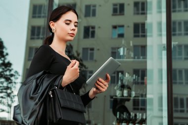 Pretty brunette caucasian woman dressed leather suit with shoulder bag walking outside hold tablet pc, order taxi, video chat call, typing message texting chatting, search information on the Internet