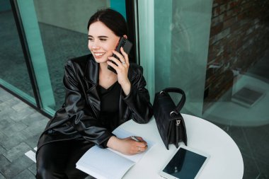 Pretty brunette caucasian woman dressed leather suit with shoulder bag sitting outside cafe street speaking on phone, writing notes. Businesswoman smiling lady. Professional business owner