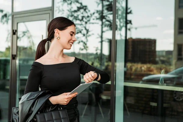 Pretty brunette caucasian woman dressed leather suit with shoulder bag stand outside hold tablet pc, looking at swart wrist wireless watch fitness bracelet, checking time, waiting in a hurry, punctual