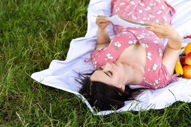 Woman lying on white bedsheet, hold and read book notes. Lady have a rest in the city park, green grass lawn. A lot of fruits around. Travel, vacation, picnic set