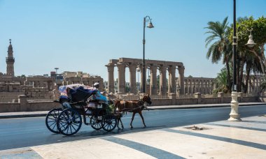 Luxor, Mısır - 27.07.2025. Geleneksel bir at arabası Luxor Tapınağı 'nın antik kalıntılarını ve açık mavi gökyüzünün altındaki bir şehir caddesindeki camiyi geçiyor.