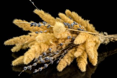 Kurumuş altın çimenler ve lavanta dallarından oluşan kırsal bir buketin yakın plan görüntüsü yansıtıcı siyah bir yüzey üzerinde ustalıkla düzenlenmiştir.