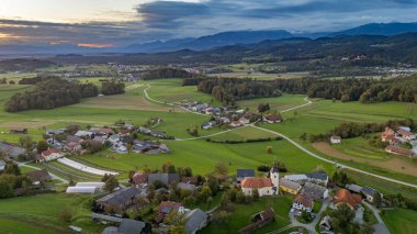 Slovenya 'nın Lukovica kenti yakınlarındaki Spodnje Koseze' nin hava manzarası, yeşil tarlaları, kırmızı çatılı evleri ve dramatik bir gökyüzü altında ormanlık tepeleri gösteriyor.
