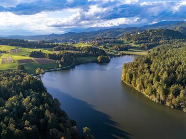 Slovenya 'nın Lukovica kenti yakınlarındaki Gradisko Jezero' nun hava görüntüsü, öğleden sonra saatlerinde verimli ormanlarla çevrili sakin bir gölü ve parçalı bulutlu bir gökyüzünün altında yuvarlanan tepeleri gösteriyor.