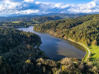 Slovenya 'nın Lukovica kenti yakınlarındaki Gradisko Jezero' nun hava görüntüsü, öğleden sonra saatlerinde verimli ormanlarla çevrili sakin bir gölü ve parçalı bulutlu bir gökyüzünün altında yuvarlanan tepeleri gösteriyor.