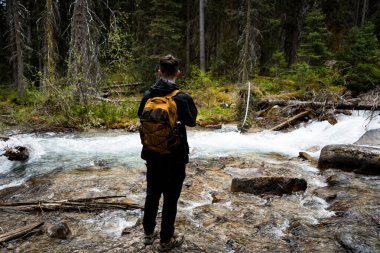 Ormanda nehrin kenarında duran, sırt çantası takan bir adam.