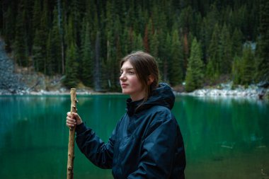Açık hava kıyafetleri içinde genç bir kadın zümrüt gölünün yanında sopayla dinleniyor.