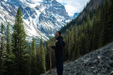 Dişi yürüyüşçü, arka planda karla kaplı dağlarla kayalık yolda duruyor.
