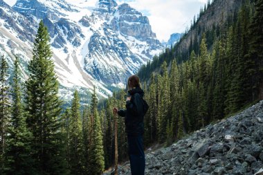 Dişi yürüyüşçü, arka planda karla kaplı dağlarla kayalık yolda duruyor.