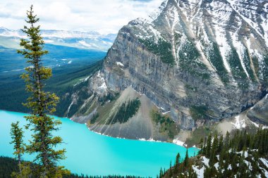 Turkuaz bir göl üzerindeki karlı uçurumlar, yeşil bir orman ve yeşil bir yamaç.