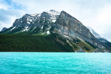 Karla kaplı kayalık dağ turkuaz göl ve sık çam ormanı üzerinde yükseliyor.