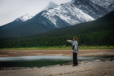 A person points toward majestic snow-dusted mountains across a lake