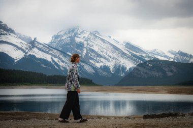 Person walking along lakeside with snow-covered mountains in background