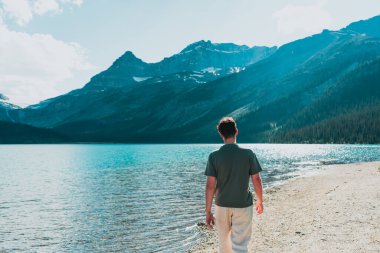 Man walking on lakeside beach with mountain range background.