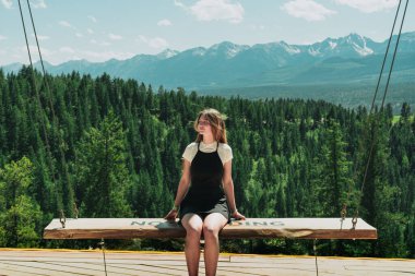 Woman on mountain swing enjoys scenic vista of forest and mountain range.