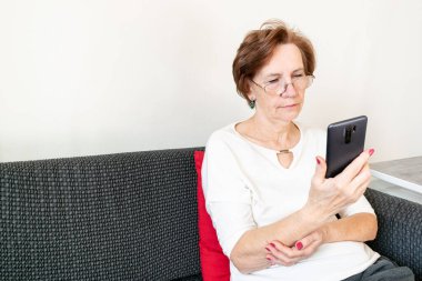 old senior woman female sit home flat couch hold smartphone write text watch video call alone unhappy sad thinking.