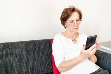 old senior woman female sit home flat couch hold smartphone write text watch video call alone unhappy sad thinking.