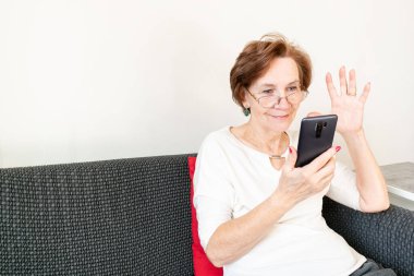 old senior woman female sit home flat couch hold smartphone write text watch video call alone happy sad thinking.