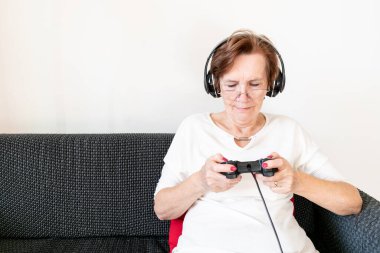 old senior woman female sit home flat couch hold joystick controller play game happy headphones alone.