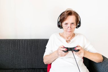 old senior woman female sit home flat couch hold joystick controller play game happy headphones alone.