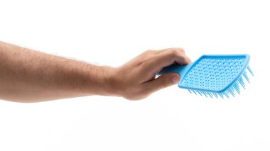 Bir el, dokulu bir yüzeyi olan mavi bir saç fırçasını kavrar ve ince kılları ortaya çıkarır. Arka plan düz beyaz, fırça ve tımarla ilgili el vurgulanıyor..