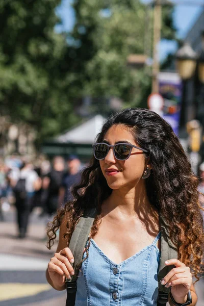 attractive young turkish tourist walking the street with a backpack and looking away. She wears a denim dress and sunglasses