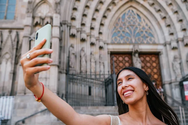 stock image Happy Latin American female tourist with long black hair smiling and taking selfie against entrance of old cathedral in Barcelona, Spain