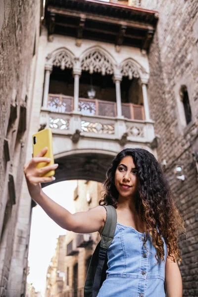Young turkish female tourist with long curly hair taking selfie at Barcelona old town streets