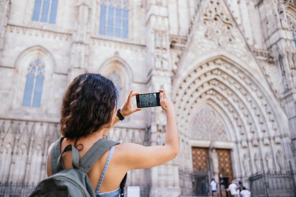 年轻的黑发姑娘背着背包拿着智能手机给巴塞罗那大教堂拍照在白天探索历史名城的街道 — 图库照片