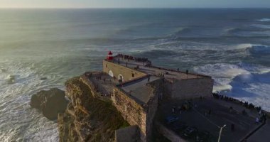 Büyük bir uçurumun üzerindeki deniz fenerinin havadan görünüşü ve Portekiz, Nazare 'nin devasa okyanus dalgalarının görüntüsünü sunuyor..