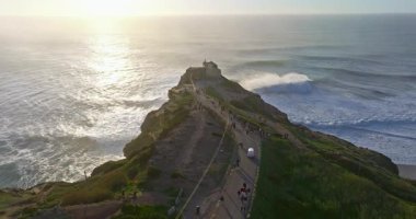 Büyük bir uçurumun üzerindeki deniz fenerinin havadan görünüşü ve Portekiz, Nazare 'nin devasa okyanus dalgalarının görüntüsünü sunuyor..