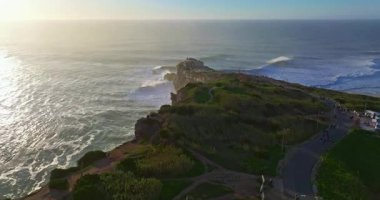 Büyük bir uçurumun üzerindeki deniz fenerinin havadan görünüşü ve Portekiz, Nazare 'nin devasa okyanus dalgalarının görüntüsünü sunuyor..
