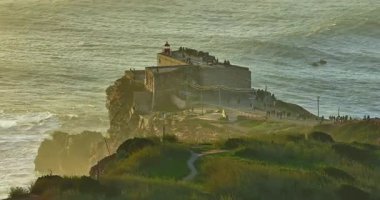 Büyük bir uçurumun üzerindeki deniz fenerinin havadan görünüşü ve Portekiz, Nazare 'nin devasa okyanus dalgalarının görüntüsünü sunuyor..