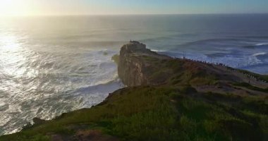 Büyük bir uçurumun üzerindeki deniz fenerinin havadan görünüşü ve Portekiz, Nazare 'nin devasa okyanus dalgalarının görüntüsünü sunuyor..