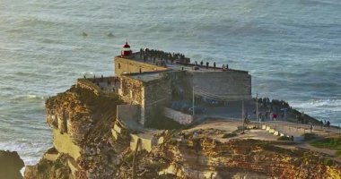 Büyük bir uçurumun üzerindeki deniz fenerinin havadan görünüşü ve Portekiz, Nazare 'nin devasa okyanus dalgalarının görüntüsünü sunuyor..