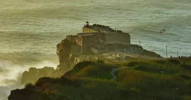 Büyük bir uçurumun üzerindeki deniz fenerinin havadan görünüşü ve Portekiz, Nazare 'nin devasa okyanus dalgalarının görüntüsünü sunuyor..