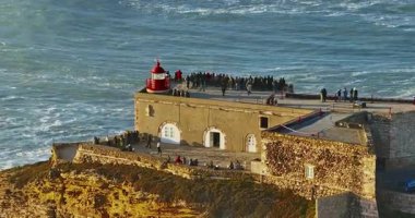 Büyük bir uçurumun üzerindeki deniz fenerinin havadan görünüşü ve Portekiz, Nazare 'nin devasa okyanus dalgalarının görüntüsünü sunuyor..