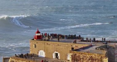 Büyük bir uçurumun üzerindeki deniz fenerinin havadan görünüşü ve Portekiz, Nazare 'nin devasa okyanus dalgalarının görüntüsünü sunuyor..