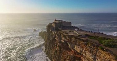 Büyük bir uçurumun üzerindeki deniz fenerinin havadan görünüşü ve Portekiz, Nazare 'nin devasa okyanus dalgalarının görüntüsünü sunuyor..